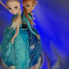 A close-up of the Anna and Elsa doll cake, showing the intricate buttercream dress details against a dramatic blue light.