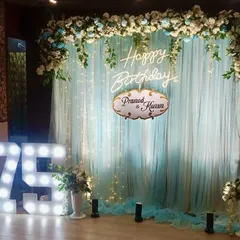An elegant 75th birthday celebration with a pastel blue and white theme. The backdrop features soft blue drapes with fairy lights, a floral arch, and large marquee numbers.