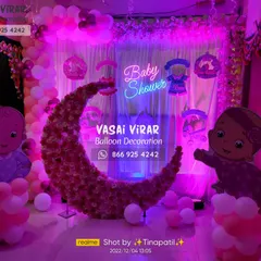 This baby shower decoration features a beautiful floral moon backdrop, perfect for photos. The setup is lit with a soft purple light and includes a "Baby Shower" neon sign and cute baby cartoon cutouts to complete the theme.