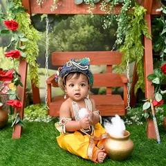 A divine moment with our little Krishna, sitting by a swing in a lush, green garden setting. The details, from the peacock feather crown to the pot of butter, make this a truly special portrait.