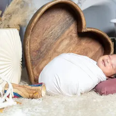 This newborn is smiling in his sleep, wrapped snugly and resting against a wooden heart prop.