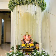This elegant Ganpati mandap features a canopy of cascading white flowers, creating a divine and pure ambiance. The design is complemented by lush white and green floral arrangements at the base, making it a serene centerpiece for your home celebration.