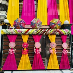 A close-up of a home's gate decorated for a Mehendi or wedding. The bright pink and yellow drapes are accented with flowers and traditional Rajasthani umbrellas.