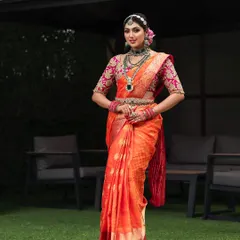 An outdoor bridal portrait where the vibrant orange and pink saree stands out against the natural green background. This shot showcases a complete bridal look, from the diamond matha patti to the embroidered blouse.