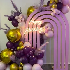 A trendy and glamorous birthday setup featuring a pink rainbow arch prop. The decor is elevated with a "Happy Birthday" neon sign and a rich balloon arrangement in shades of purple, pink, and gold.