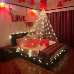 This romantic room decoration is bathed in a warm red glow. The setup includes an illuminated canopy, "I Love You" foil balloons, and rose petals on the floor for a passionate and memorable surprise.