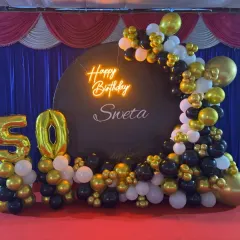 Celebrate a 50th birthday in style with our custom party backdrops. This setup includes a personalized circular backdrop with a neon 'Happy Birthday' sign, a large '50' marquee number display, and a sophisticated black, gold, and white organic balloon arch.