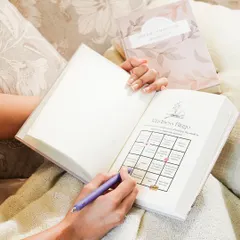 A lifestyle shot showing someone filling out a "Kindness Bingo" in their journal while relaxing on a couch. This image connects the product with positive, mindful activities and self-improvement.
