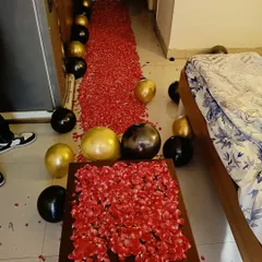 A romantic room setup featuring a long pathway of red rose petals leading to the bed, with black and gold balloons scattered around.