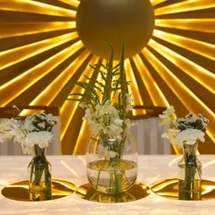A modern and glamorous tablescape for a Roka ceremony. We used simple floral arrangements in glass vases on gold placemats, set against a stunning backlit gold sunburst backdrop.