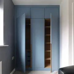 A modern, floor-to-ceiling wardrobe with a matte blue finish and long wooden handles. The doors open to reveal a well-organized interior with shelves, drawers, and hanging space.