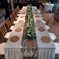 For a formal corporate dinner, we designed this elegant long-table setup. The decor features a silver sequin table runner, a lush green and white floral centerpiece, woven placemats, and candle lanterns for a warm, inviting feel.