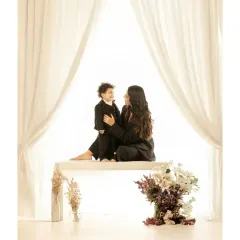 A tender moment between a mother and child in a bright, airy studio setting. Dressed in matching black outfits, they share a quiet look, framed by soft white curtains that create a feeling of intimacy and peace.