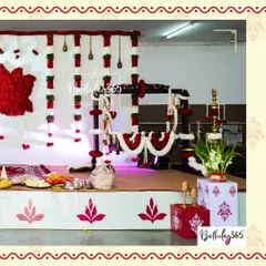 An elegant and traditional Namakaran (naming ceremony) setup. The centerpiece is a beautifully decorated wooden cradle, or 'jhoola', adorned with garlands of red and white flowers, set against a backdrop of floral hangings and brass bells.