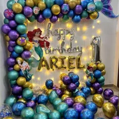 For this first birthday, I designed a complete Little Mermaid theme. The setup includes a custom "ARIEL" name foil, character cutouts, and a vibrant balloon garland in mermaid-inspired colors of purple, teal, and gold, all lit by warm fairy lights.