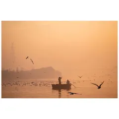 A boatman and passenger on the Yamuna River at sunrise, surrounded by a flock of migratory birds. The hazy, golden light creates a dreamy and picturesque scene.