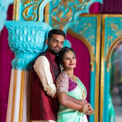A beautifully dressed couple in traditional Indian wedding attire poses against a vibrant, ornate backdrop, perfect for a reception or sangeet look.