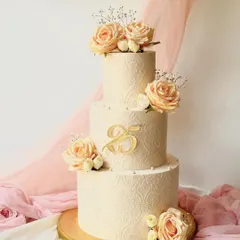 A classic and timeless three-tier anniversary cake with an intricate white-on-white stencil pattern. It's adorned with delicate peach roses and a custom gold "25" topper, perfect for a silver jubilee.