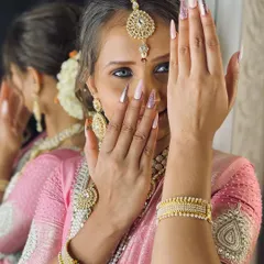 A beautiful shot of a bride showcasing her long, elegant nail extensions. The design is a soft, shimmery pink that perfectly matches her traditional pink and gold saree.