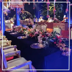 We design dining experiences that match the mood of your celebration. This image captures a luxurious sit-down dinner setting for a sangeet night, featuring a long table dressed in black velvet, accented with silver charger plates and a magnificent floral runner.