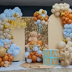 A beautiful and modern "Oh Baby" shower setup. This decoration features a neutral color palette with blue, brown, and beige balloons, a large teddy bear, "BABY" blocks, and a sequin backdrop for a touch of sparkle.