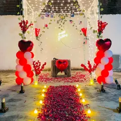 An intimate and romantic outdoor cabana setup, perfect for a birthday or anniversary surprise. The decor includes a candle-lit rose petal pathway, balloon pillars, and a canopy decorated with fairy lights and a 'Happy Birthday' neon sign.