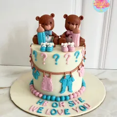 He or she? We love you! This adorable gender reveal cake features two teddy bears, one with a pink bow and one with a blue bow, surrounded by tiny baby clothes.