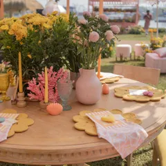 A close-up of the pastel-themed table setting. Yellow flower-shaped placemats, pink coral decor, and colorful candles create a playful and chic tablescape.