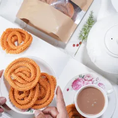 Hands sharing a plate of my Garlic Murukku with a cup of chai. My snacks are meant to be shared, bringing people together over good food.