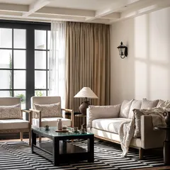 Another view of the 'Riviera Maison' living room, where natural light floods in through large, black-framed French doors. The arrangement of the furniture invites conversation, embodying the easy elegance of a European home.