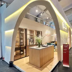 A full view of the Plüsch booth, showcasing the arched entrance and the curated interior. The design uses a monochromatic bouclé facade and soft, integrated lighting to create a warm and sophisticated skin that envelops the space.
