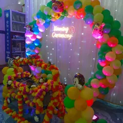 A jungle-themed naming ceremony with a beautifully decorated cradle (palna) covered in marigold flowers. The backdrop is a vibrant green and yellow balloon circle with a custom neon sign.
