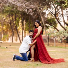A candid and joyful moment during an outdoor maternity shoot. The partner hugs the baby bump from a kneeling position, both sharing a smile under a blossoming tree.