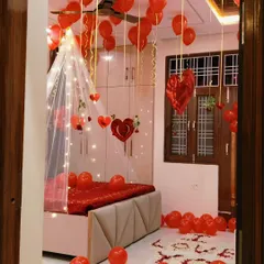A beautiful and romantic room setup, viewed from the doorway to maximize the surprise. The decor features a lit canopy, red balloons, and 'I LOVE YOU' spelled out with petals on the floor.