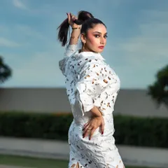 A high-fashion outdoor portrait for a magazine. The pose and high ponytail hairstyle complement the structured, modern design of the white laser-cut outfit.