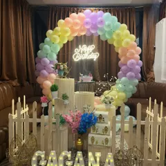 A birthday party setup featuring a full ring balloon arch in pastel rainbow colors. The decor is accented with a neon "Happy Birthday" sign, a small picket fence, and marquee letters spelling the name in front.