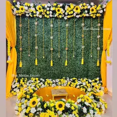 A grand Haldi setup ideal for home or banquet spaces. The design is built around a lush green wall, a floor-level urli surrounded by sunflowers and white florals, and bright yellow drapes to complete the look.
