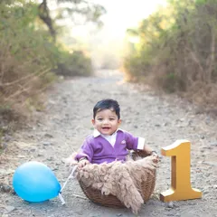 A happy one year old sitting in a basket on a rustic path. The warm, golden hour sunlight adds a magical glow to this outdoor milestone portrait.
