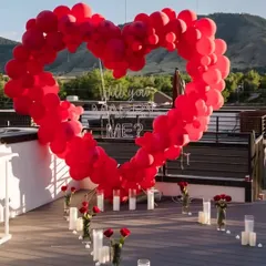 A stunning rooftop proposal setup waiting for the special moment. The focal point is a large heart-shaped arch made of red balloons framing a "Will you marry me?" neon sign against the evening sky.