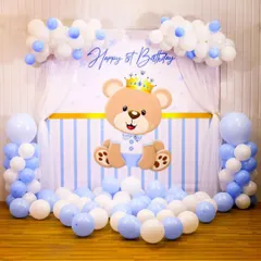 A sweet teddy bear theme for a first birthday, perfect for a baby boy. The backdrop features a cute crowned bear, with balloon columns and clouds in a soft blue and white palette.