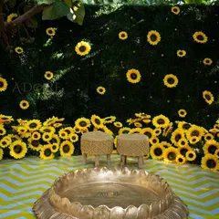 This sunflower-themed Haldi setup is designed for happiness. It features a backdrop of sunflowers on a green wall, a large brass 'uruli' for the ceremony, and a cheerful yellow-and-white chevron floor.