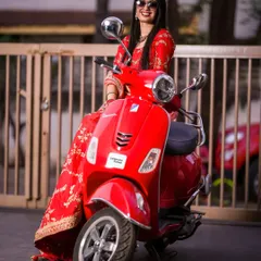 A cool bride in a red lehenga and sunglasses, posing confidently on a vibrant red Vespa. A perfect mix of tradition and modern swag.