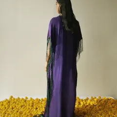 A quiet, contemplative moment from an editorial shoot. The styling focuses on the silhouette of a purple kaftan with green fringe, set against a bed of yellow marigolds, creating a rich color story with a minimalist composition.