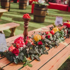 A rustic picnic table transformed with a stunning runner of fresh vegetables and flowers for a 'La Tomatina' themed Haldi. This centerpiece features red and yellow bell peppers, pomegranates, and roses, showcasing a creative and sustainable approach to event decor.