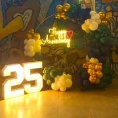 A 25th-anniversary celebration setup with large, light-up marquee numbers. The backdrop features a 'Happy Anniversary' neon sign and a colorful balloon arrangement against a graffiti wall for a modern, edgy look.