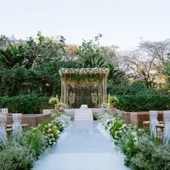 A pristine white aisle lined with lush white and green florals leads to a beautiful mandap nestled in a grove of trees. This design creates a serene, natural cathedral for the wedding ceremony.