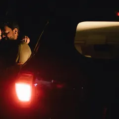 A quiet, intimate moment in the back of a car at night. The red taillight casts a soft glow on the couple as they embrace, creating a feeling of being in their own private world.