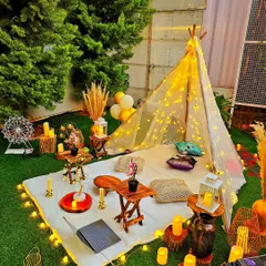 A close-up of a beautifully styled boho picnic at night. The setup includes a teepee tent wrapped in fairy lights, low wooden tables, plush cushions, and an array of candles and lanterns for a warm and inviting atmosphere.