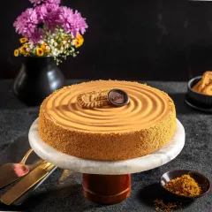 A luxurious Biscoff cheesecake on a marble stand, shot with moody, dramatic lighting that highlights its smooth texture and rich caramel tones for Bakingo.