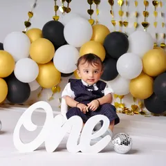 A chic and modern first birthday theme featuring a black, white, and gold balloon wall. The little gentleman in his vest looks ready to celebrate his big day in style.